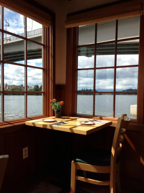 A small, wooden, corner table with one chair is situated in a windowed corner that looks out over the Port Washington Narrows and the Narrows Bridge in Bremerton, WA.