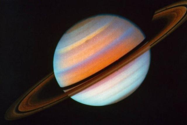 A false-color image of Saturn taken by Voyager 1 in 1980. The planet's cloud bands appear as orange, yellow, white, and a pale blue-green. The ring system is a darker golden-brown, and the portion circling behind the planet is partly in shadow. The image is tilted at a roughly 30 degree angle, with the equatorial ring system stretching from the lower left to upper right corners of the photo.
