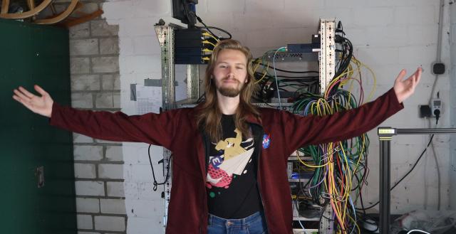 a picture of Erik Uden wearing the newest and first mastodon T-Shirt, above that a NASA jacket, also wearing a blue jeans. Behind him there is a server rack with many cables, networking devices, and servers. On the bottom right you can see a 3D printer. The whole setting is inside of a garage. 