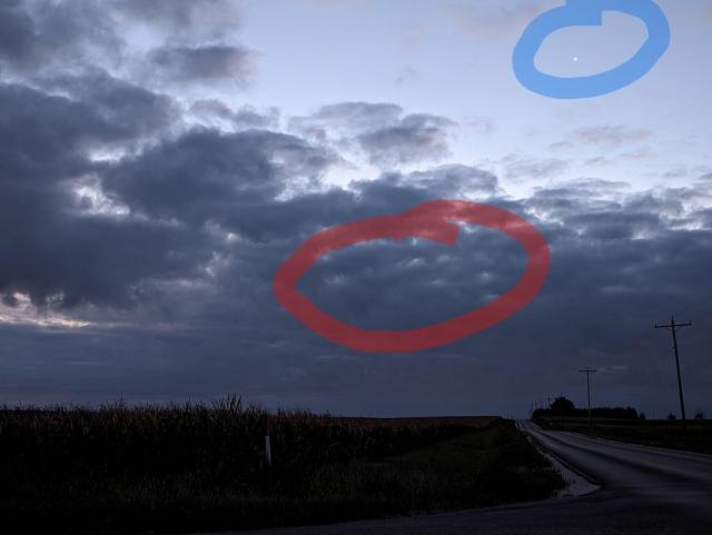 Dawn breaking over the country side with dark corn fields below and a lightening sky above. There are dark clouds up through the horizon and stretching quite far up into the sky. Two circles are drawn, blue one around a tiny prick of light and red one around a section of the dark clouds. 
