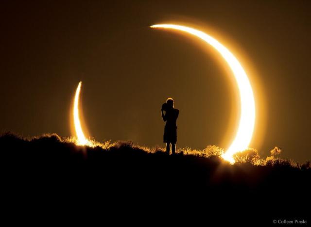 A person is seen standing at the top of a ridge. The person appears as a silhouette onto the central dark region of an annular solar eclipse. The annular solar eclipse is a bright ring with a large dark hole in the middle.