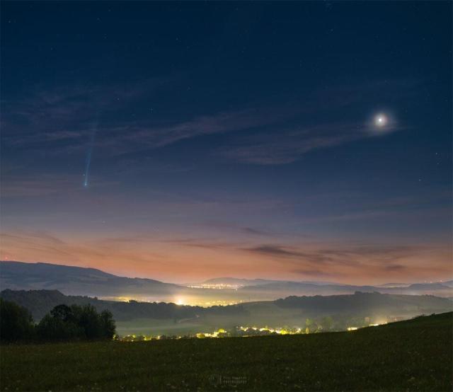 A scenic and hilly landscape is shown just before sunrise. On the left is Comet Nishimura near the horizon with a long tail fading off toward the top of the frame. On the right is a bright spot that is Venus. The sunrise sky is dark blue at the top but morphs into tan at the horizon, while the foreground hills are green.