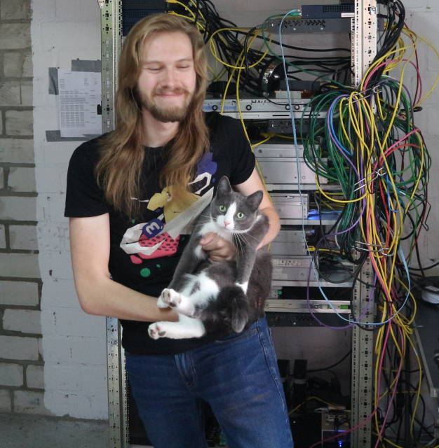 A picture of me waring the official Mastodon T-Shirt carrying my cat Kitty and smiling like a sweet pickle. The mastodon.de servers can be seen in their monstrosity in the background.
