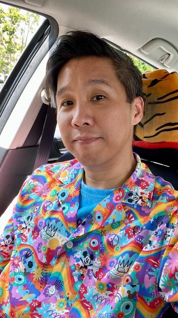 Asian guy facing the camera with a slight smile. His shirt features Pride rainbow pattern all over, with Mickey Mouse and Minnie sprinkled throughout wearing Pride rainbow outfits. 