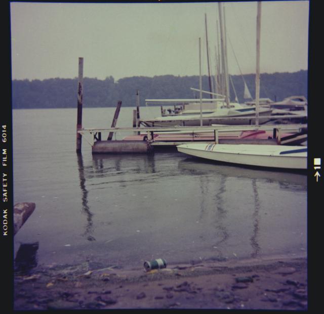 photo of a color film shot of some small boats by a shore. Treeline is in the distance indicating this might be a lake. Discarded can by the water.