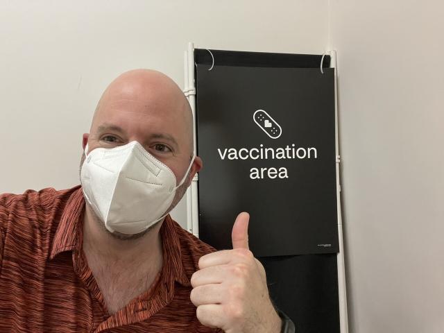 Jon Masters sitting in a vaccination area with a sign saying “vaccination area” to his left
