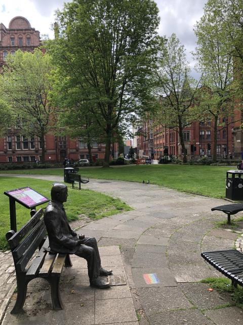 Side profile of Turing memorial. Made of solid cast brass or something, with him sitting on a bench. In the middle of a park in a big city. 