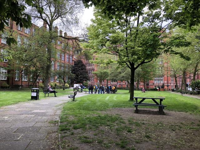 Zoomed out view of park. Four story building surround a small square park that has some grass and trees and a silent spot with the memorial. 