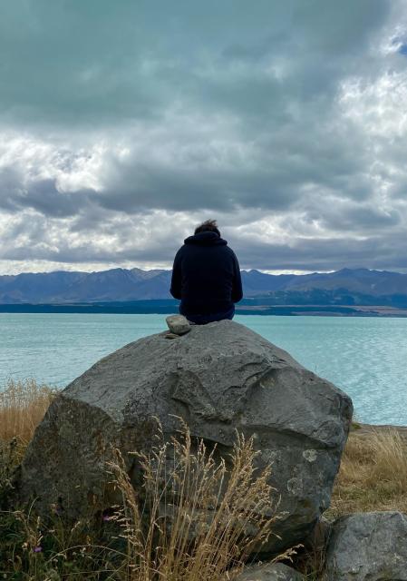 Landschaftsfoto mit einem großen Felsbrocken an einem See (Lake Tekapo/NZ) und Bergen im Hintergrund. Auf dem Stein sitzt ein Mensch im Kapuzenpulli mit vom Wind zerzaustem Haar. Man sieht ihn von hinten. Die Wolken hängen tief.