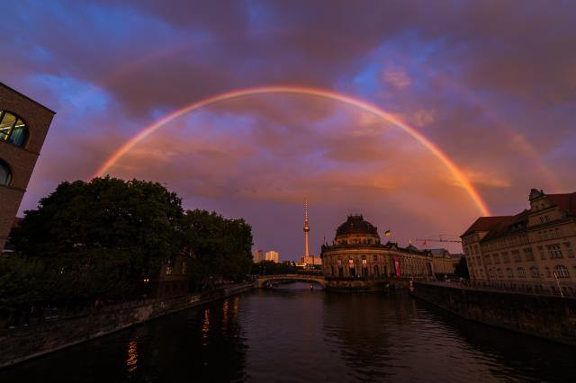A very bright primary rainbow and a fainter second bow arc high above the Berlin skyline, with the Museumsinsel (Museum Island) in the foreground, and Fernsehturm (TV tower) behind