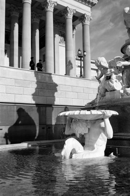 Black and white picture of the Austrian parliament. The fountain is casting a shadow on the outside of the building. On top of the shadow next to some pillars are two policemen.