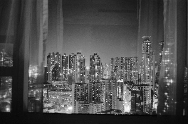 Black and white photo of the Mong Kok city skyline at night through a window framed by diaphanous curtains. 