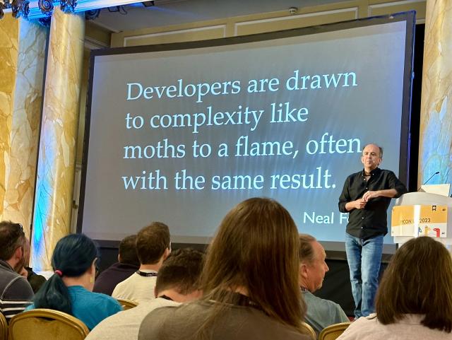 Kevlin Henny on stage in front of a slide with a quote that says “Developers are drawn to complexity like moths to a flame, often with the same result.”