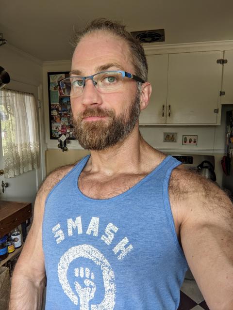 Bearded white guy stands in a kitchen wearing a tired expression and a blue tank top with the word SMASH on it.