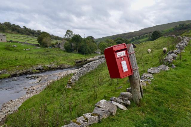 A bright red postbox on a post in the foreground, a couple of stone walls looking slightly the worse for wear and a couple of sheep grazing in an open field. There's a stream running through the centre of the picture, through a green valley of walled fields and barns. The sky is slightly overcast.