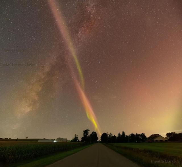 A rural road is pictured running to the horizon with rural grassy fields on both sides. Rising from the lower left is the central band of our Milky Way Galaxy. Rising from the horizon -- just at the visible end of the road, is a thin twisting band of light twisting green and red bands -- a STEVE. The STEVE crosses in front of the Milky Way band making a big .