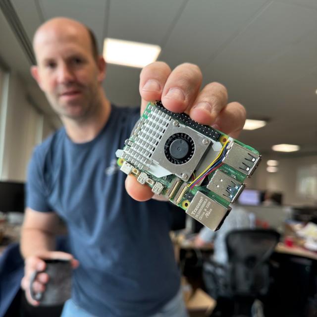 Eben stood over his desk at Pi Towers holding a Pi 5 with the active cooler attached close up to the camera