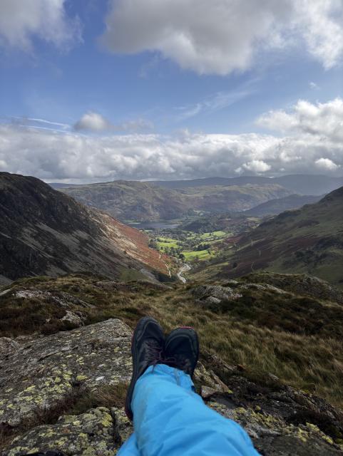 A view down in Glenridding in the UK Lake District, the weather is mixed but will sunny spells.  A pair of feet is seen reclining in the lower part of the frame.  The view is glorious over the distant fells.