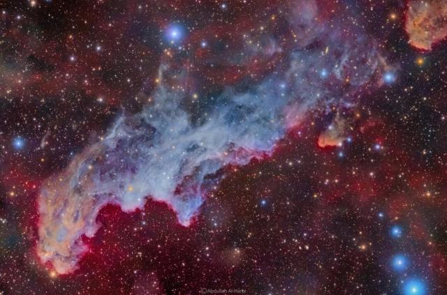 A colorful star field surrounds a big blue reflection nebula. The nebula is elongated across the wide frame and said to resemble the head of folklore-based witch.