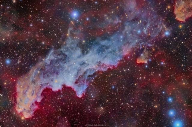 A colorful star field surrounds a big blue reflection nebula. The nebula is elongated across the wide frame and said to resemble the head of folklore-based witch.