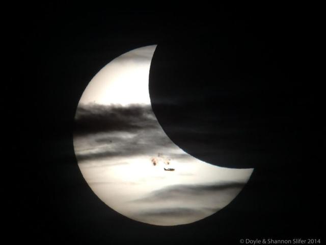 A partially eclipse Sun is shown. In front of the Sun are sunspots, the Moon, clouds, and an airplane.