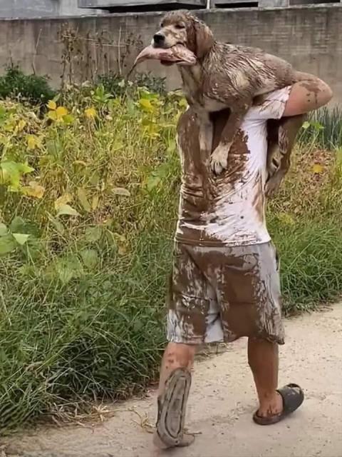A man covered in mud is carrying a muddy dog on his shoulder. A dog has a fish in its mouth. 