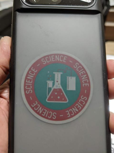 The translucent back of a phone with a round sticker with the word science repeated around the border and in the middle - a test tube, conical flask, tubing and a beaker.