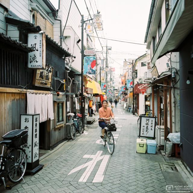 At Star Road in Asagaya, Tokyo, a few pedestrians and bicyclists are making their way between the many small shops and eateries that are typical for this area. (Daytime View)