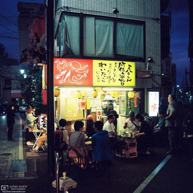On an evening outside an Izakaya (Japanese pub) in Asagaya, Tokyo, patrons are seated around small tables, and enjoying drinks and various grilled foods.