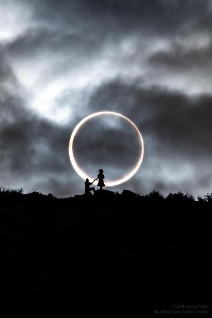 An annular solar eclipse appears in the background with the dark Moon appearing completely internal to the bright Sun. In the foreground is a ridge with the silhouettes of two people, one standing, and one kneeling.