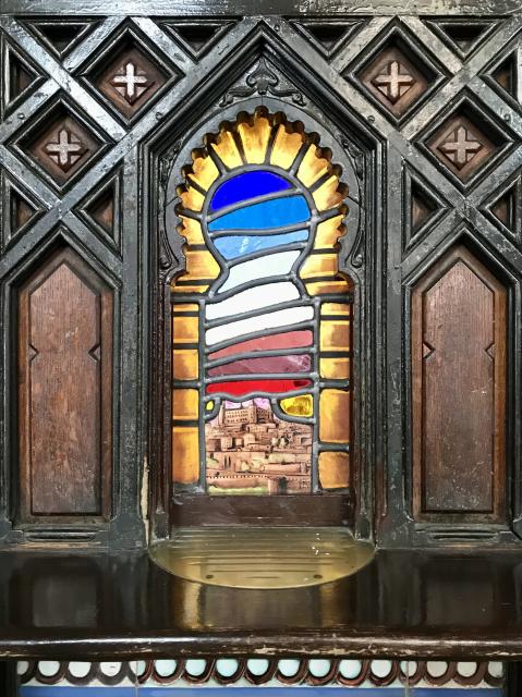 A colorful leaded window depicting the city of Toledo at dawn, with the city at the bottom and layers os sky above in red fading to dark blue to the top. The shape of the window is that of an Arab arch, and the window is surrounded by a panel of aged wood, with veins of deep brown, and adorned with a lattice of diamonds with little crosses inside. 