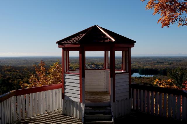 In the centre of the frame is a small cupola with a red roof on top of a mountain. Its open windows offer a wide view over the forested landscape in front of it. The forests are in orange-brown fall colours. The blue sky is a bit hazy. Close to the mountain is a small lake, and looking through the centre window a few streets and houses are visible.  
