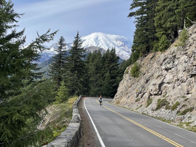 A bike rider climbs a winding road with a granite wall on one side and a short rock wall on the other.  In the background, Mt Rainier appears with a white cloud on top. 