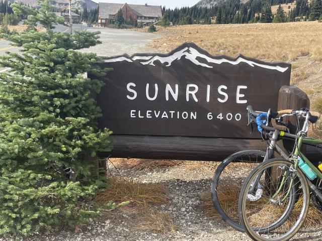 Two bikes lean across a park sign that says "Sunrise, Elevation 6400 ft"  In the background is a large empty parking lot near a lodge.