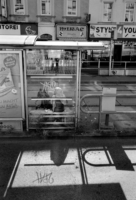 Black and white picture of a woman with a pram waiting in the tram shelter while the sun is casting a shadow of her through the glass wall.