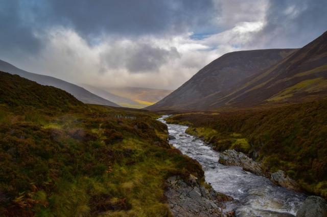 Picture of a valley in the Cairngorms in Scotland. A stream or river runs in the middle. Low clouds make some of the hills hazy though the sun breaks through in the background. The vegetation is green, yellow, orange and dark red. 