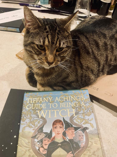 Sam (a tabby cat) sits next to an early physical copy of a Tiffany Aching’s Guide to being a Witch