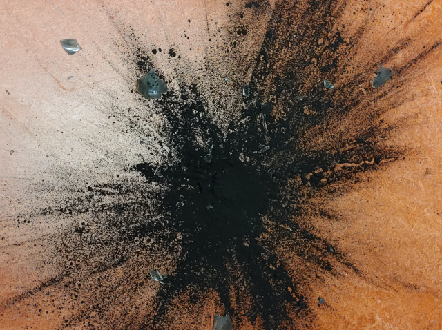 Photo of a broken glass once containing black pigment, now exploded in a star burst pattern across an orange floor.