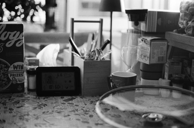A black and white photo of a dinner table with various kitchen objects on it: a box of cereal, a salt shaker, tissues, a mug, a clock, a pot lid, a cutlery holder full of spoons and knives, a sand clock, a box of glasses cleaning wipes, and more.