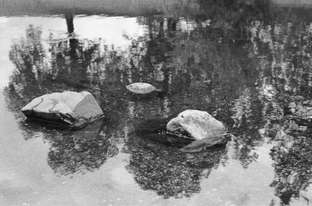 A black and white photo of 3 rocks in a body of water, with the reflection in the water revealing a line of trees.