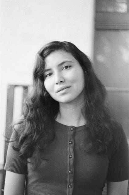 A black and white photo of a woman with dark curled hair wearing a short-sleeve button-down sweater top, with her head tilted slightly.