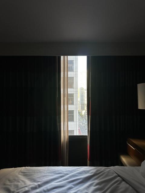 Photo with a slightly rumpled bed in the foreground, illuminated by a strip of light coming in through just slightly open curtains that take up the middle half of the shot. The edge of a high-rise building is visible in the window. 