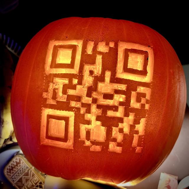 An internally lit pumpkin with a QR code carved into it encoding the text “Boo !”