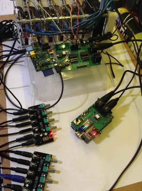 Photo showing a cluster of Raspberry Pi V1 boards at the back and a single Pi V1 with a MiniDexed user interface pcb (encoder, buttons, SSD1306 display) attached near the front.  The Pis are all joined using jumper wires and the audio outputs going into cheap PCB mixers. 