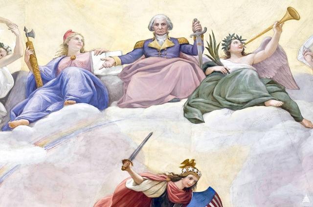 a further close up of washington, seated with one arm outstretched, and the other holding a sword. he is flanked by personified victory, a.k.a. victoria, who hold an olive branch and blows a trumpet, and personified liberty, who wears a phrygian cap and holds an open book of law and a fasces.