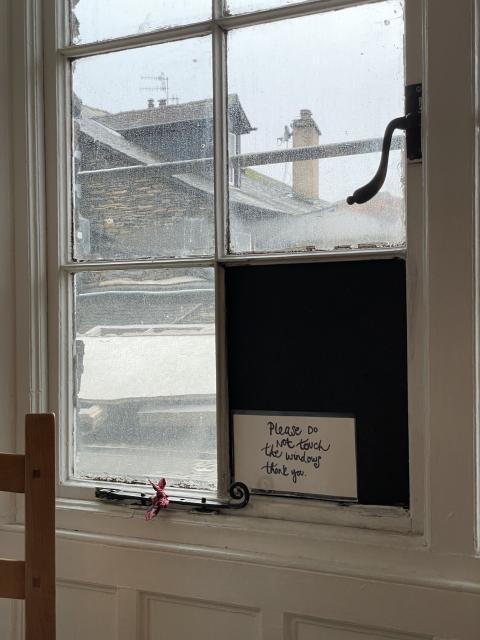 An old single glaze window covered in dust, with a white frame, looking out towards a grey stone house and roof with chimney. One of the tiles of the window is replaced with a black board, and a note on top asks in handwriting to please not touch the windows. 