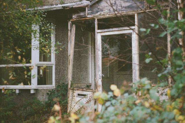 A porch entrance of an abandoned house that is slowly being overtaken by nature. The trees still have most of their leaves, but some of them are already yellow.