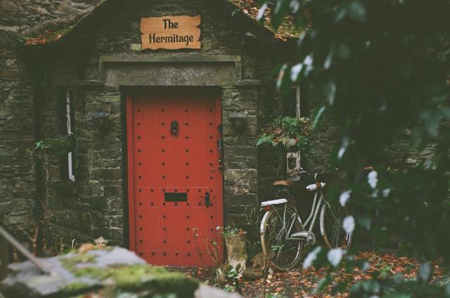 A bright red door on a corner entrance of a grey stone building with moss all over it. A bike leans on the wall next to the door. The signage above the door says "The Hermitage". 