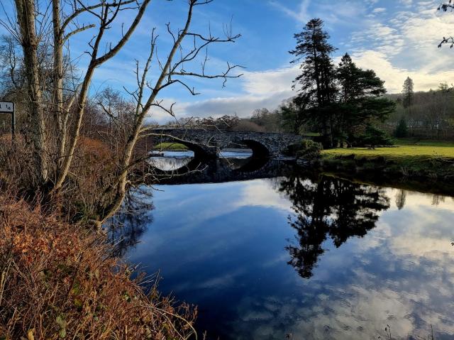 Photo of the bridge at Llanderffel in the sun with reflections in the water and autumn coloured foliage.