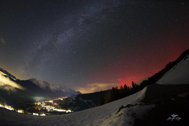 The night sky over a valley is shown complete with the central band of the Milky Way Galaxy crossing up from the lower left. On the right the sky just over the hill glows an unusual red: aurora.